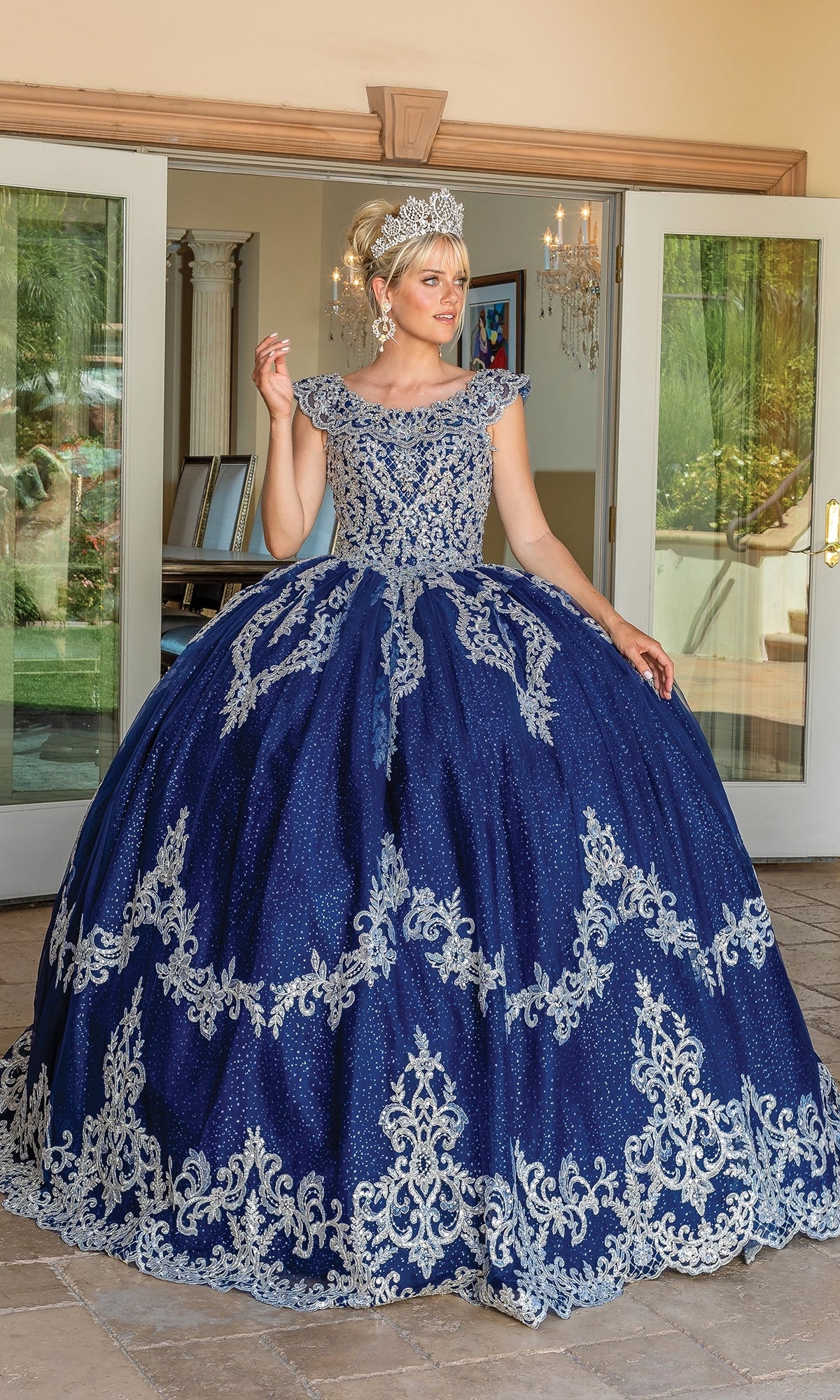 A woman in the Navy Blue Quinceanera Dress 1709 stands before open glass doors, posing elegantly with one hand raised. The gown features ornate lace embroidery and is paired with a silver tiara.